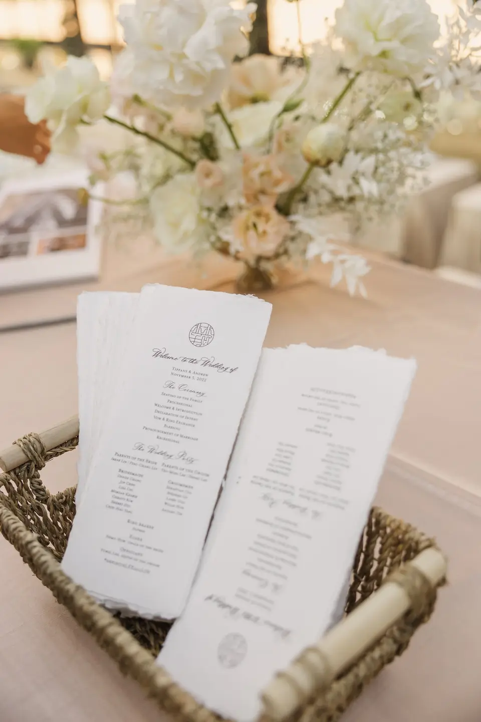 Printed ceremony programs in a woven basket on a table, beneath an arrangement of cream and blush flowers.
