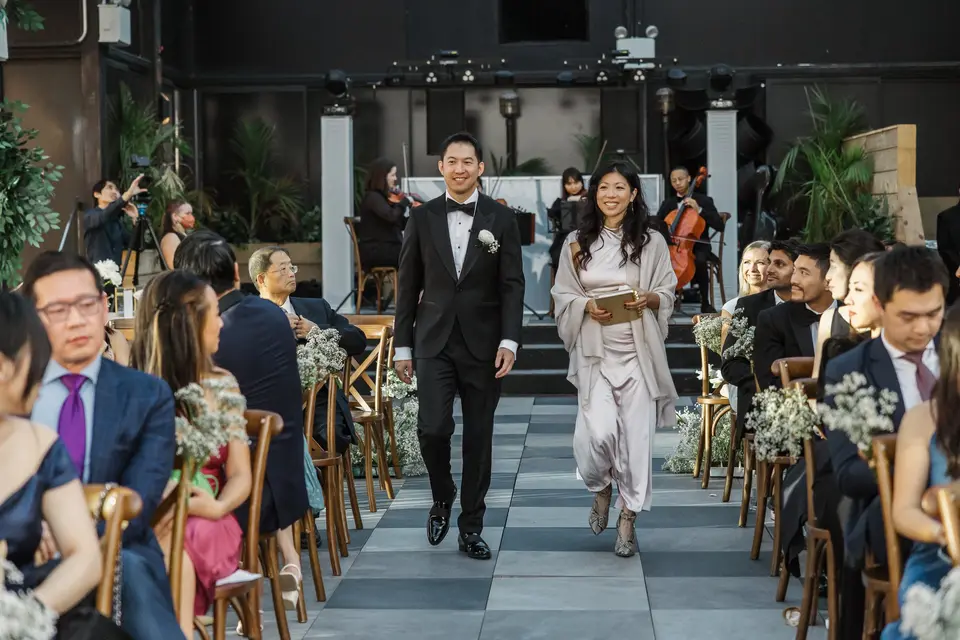 The groom walks down the checkered aisle beside a woman in blush carrying a folder, guests seated on either side.
