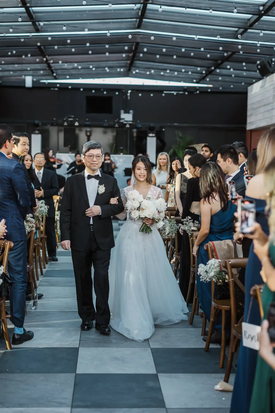 The bride walks down the aisle on her father's arm beneath string lights, guests standing on either side.