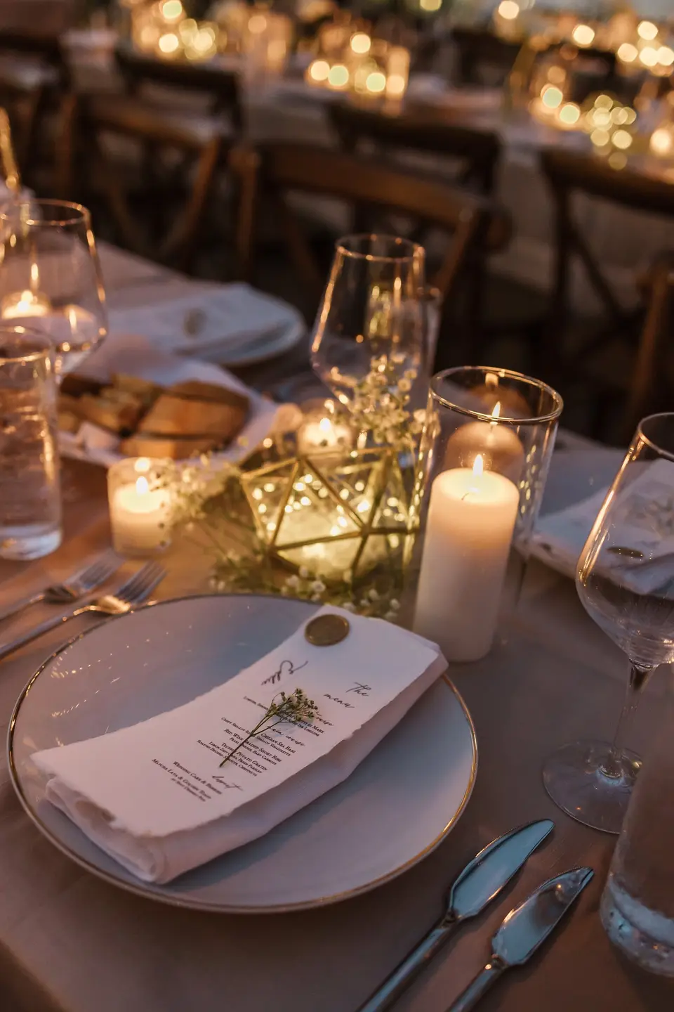A place setting with a folded menu and a sprig of greenery, pillar candles and a gold terrarium beside it.
