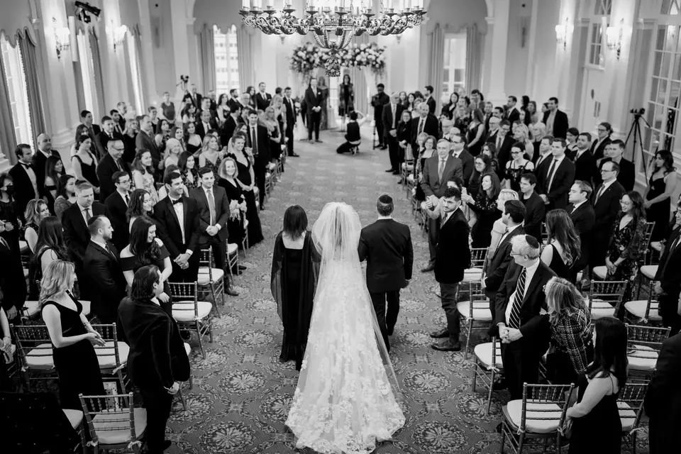 From above, the bride walks down the aisle between rows of standing guests. Black and white.