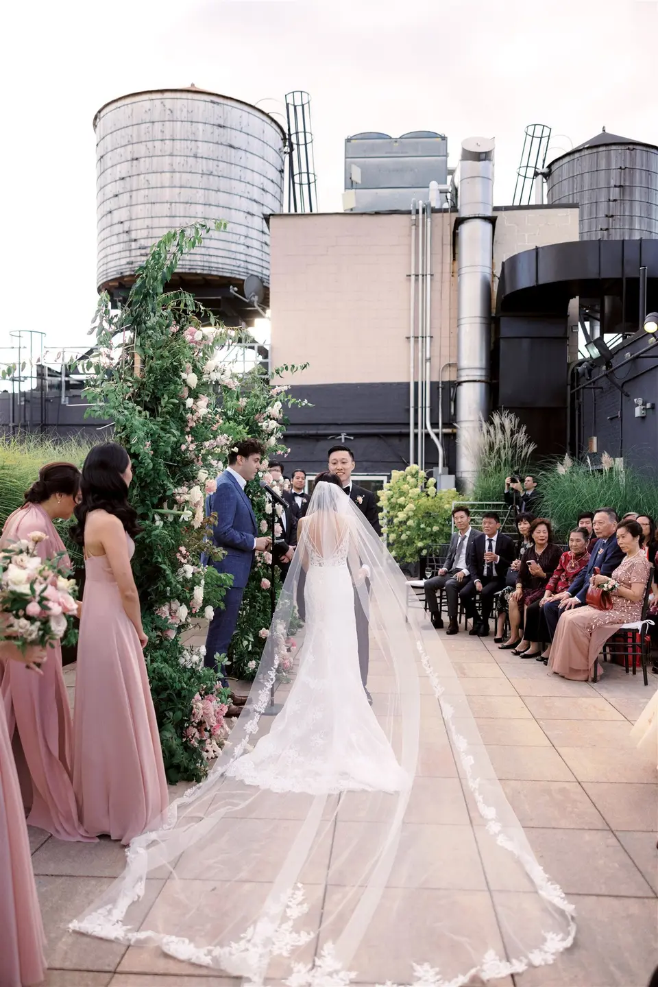 From behind, the bride stands at the arch, her cathedral veil running the length of the aisle, water towers on the roofs beyond.