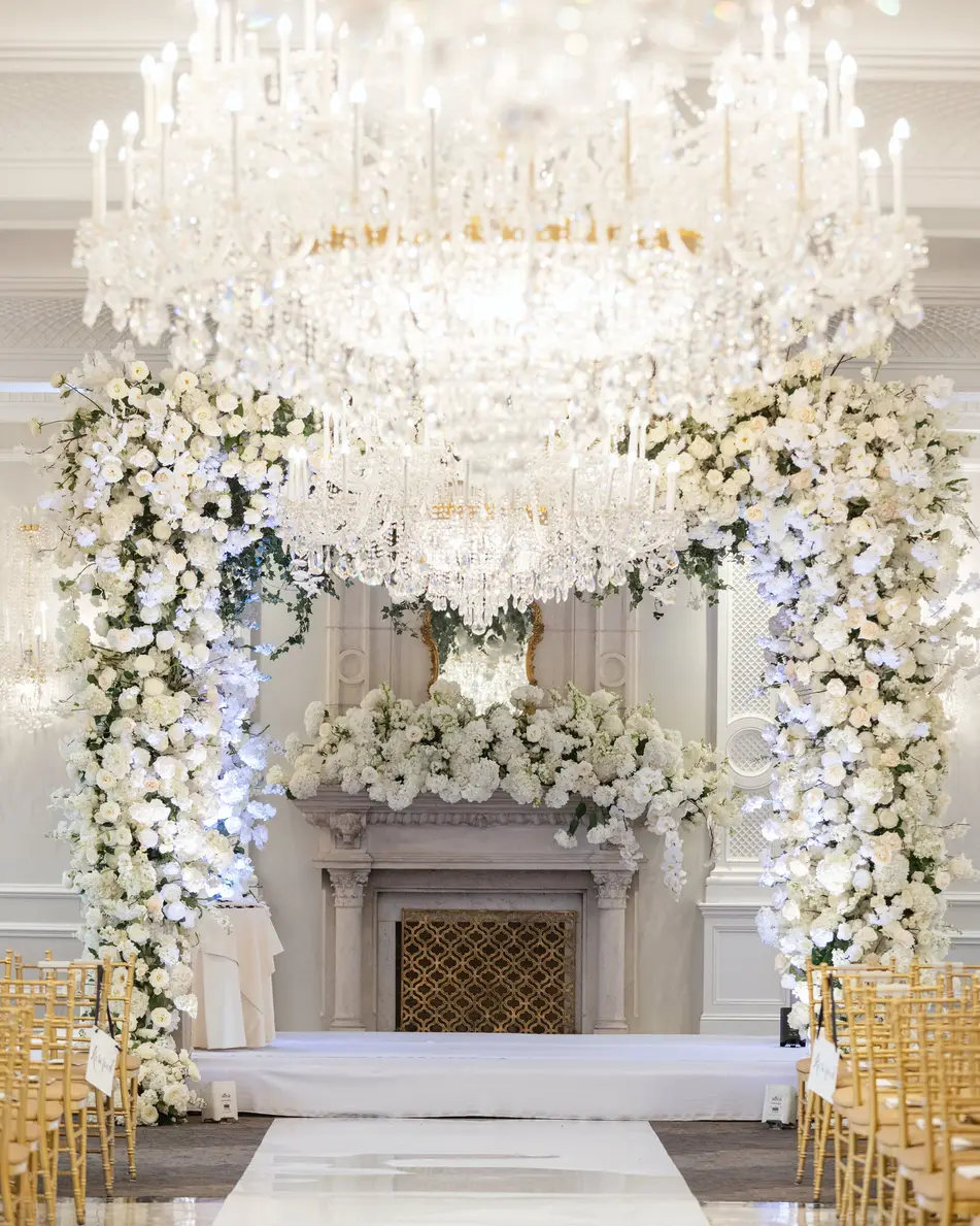 A white aisle runs to a floral arch around the fireplace, a crystal chandelier above and gold chairs on either side.