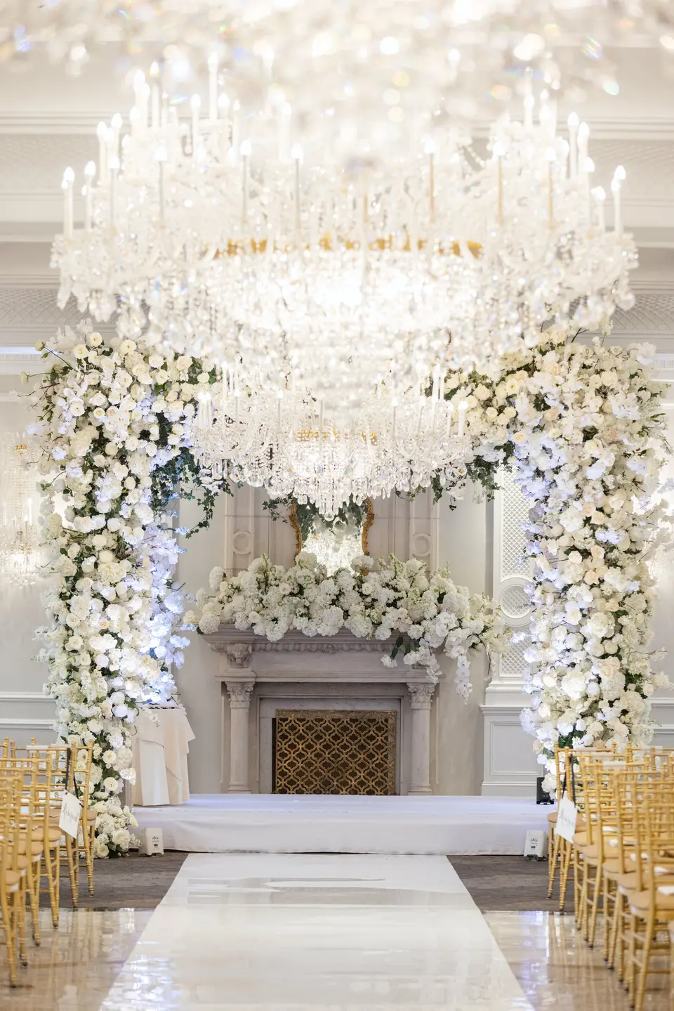 A white aisle runs to a floral arch around the fireplace, a crystal chandelier above and gold chairs on either side.