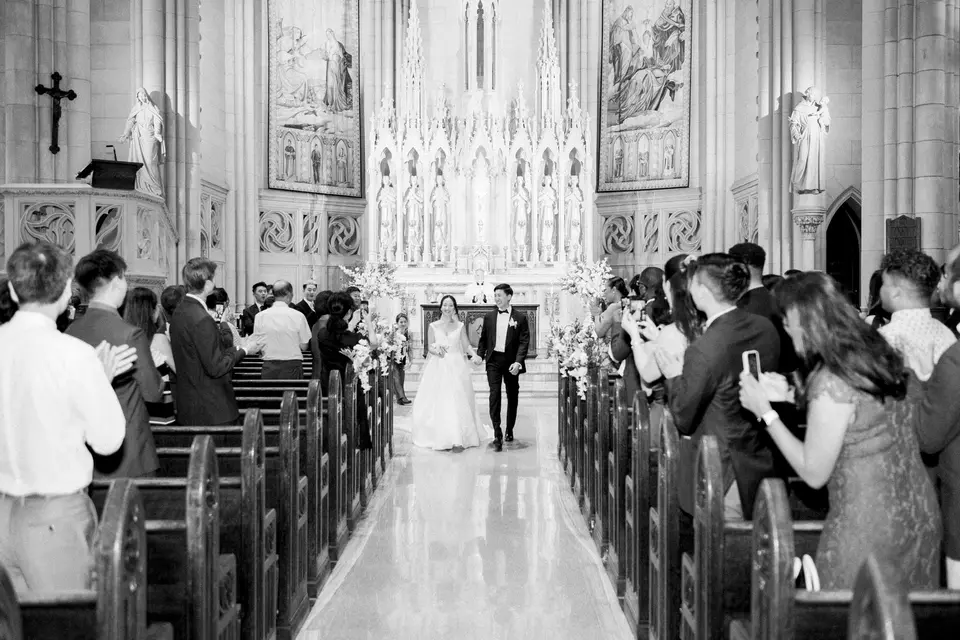 The couple walk back up the long aisle between the pews as guests stand and applaud. Black and white.