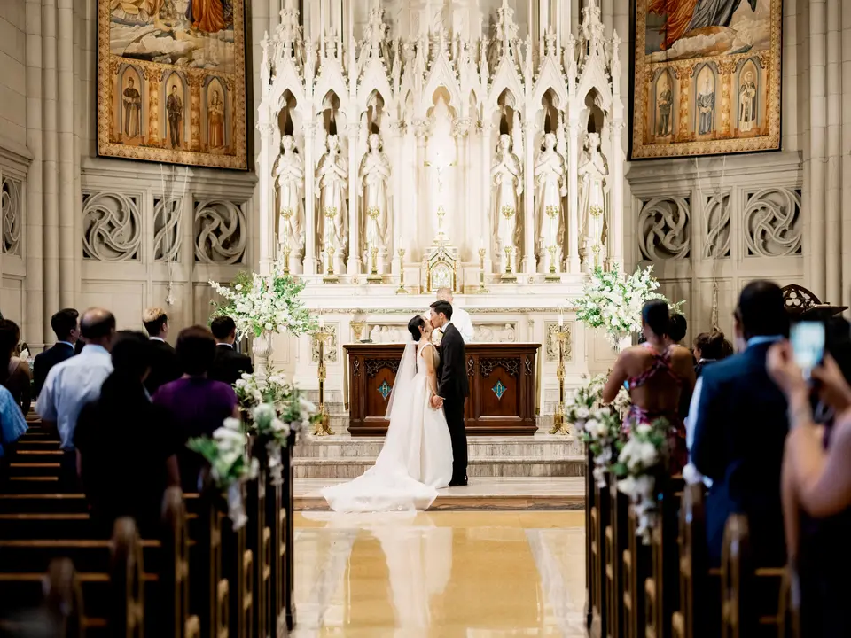The couple stand at the white marble altar beneath gilded paintings, white flowers on either side and guests in the pews.