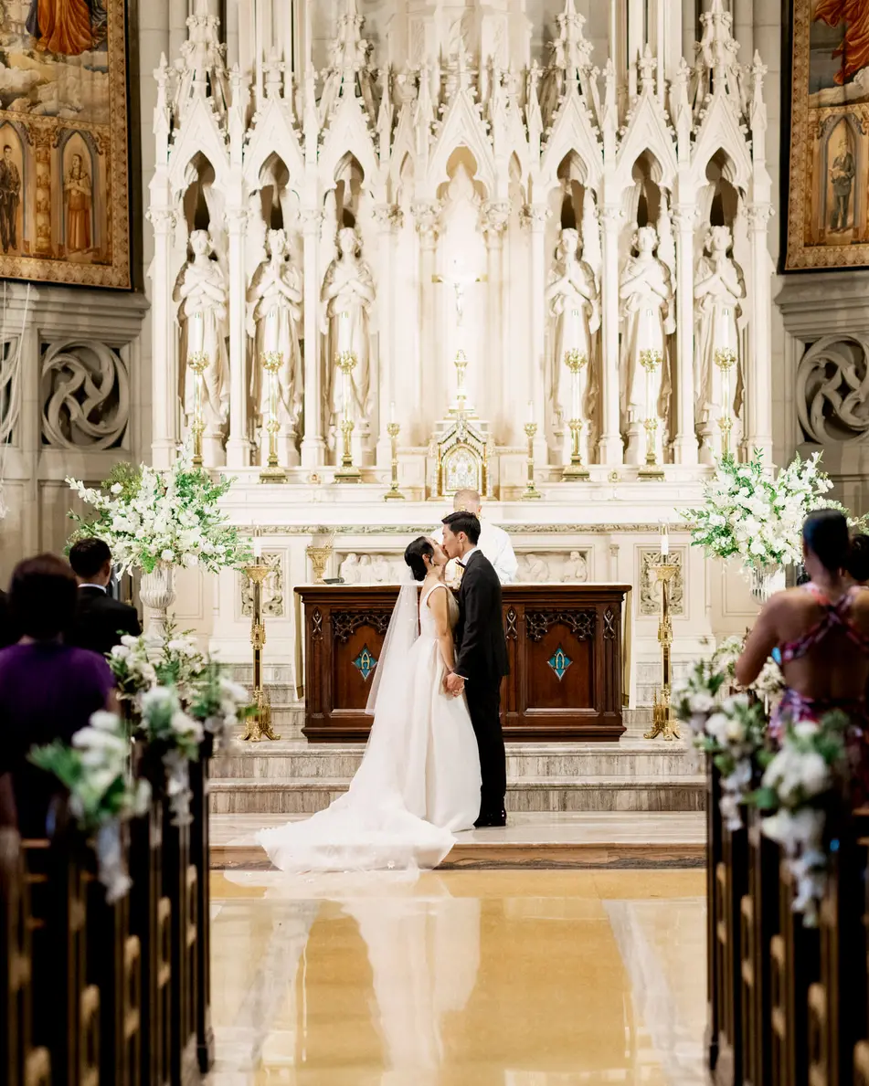 The couple stand at the white marble altar beneath gilded paintings, white flowers on either side and guests in the pews.