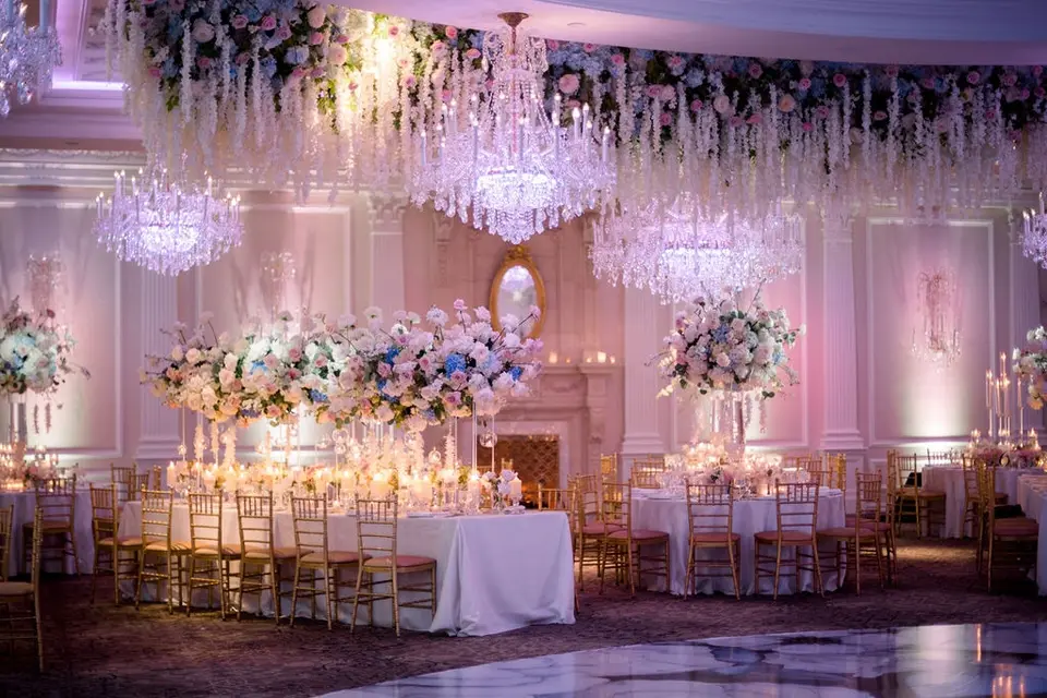 The ballroom at The Rockleigh on the night, white wisteria and flowers hanging above crystal chandeliers, long tables glowing with candles and tall blush and blue arrangements, a marble dance floor in the foreground.