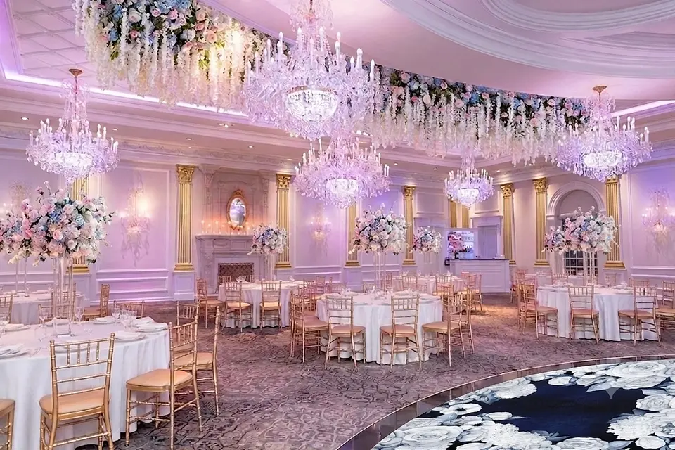 A rendering of the ballroom at The Rockleigh, flowers and white wisteria hanging above the crystal chandeliers, tall blush and blue arrangements on round tables with gold chairs, and a dance floor patterned with white roses on navy.