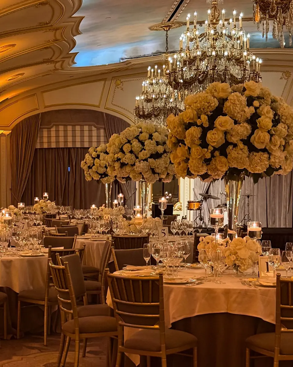 A ballroom at The St. Regis New York set for dinner, tall arrangements of cream roses on round tables beneath crystal chandeliers and a gilded vaulted ceiling.