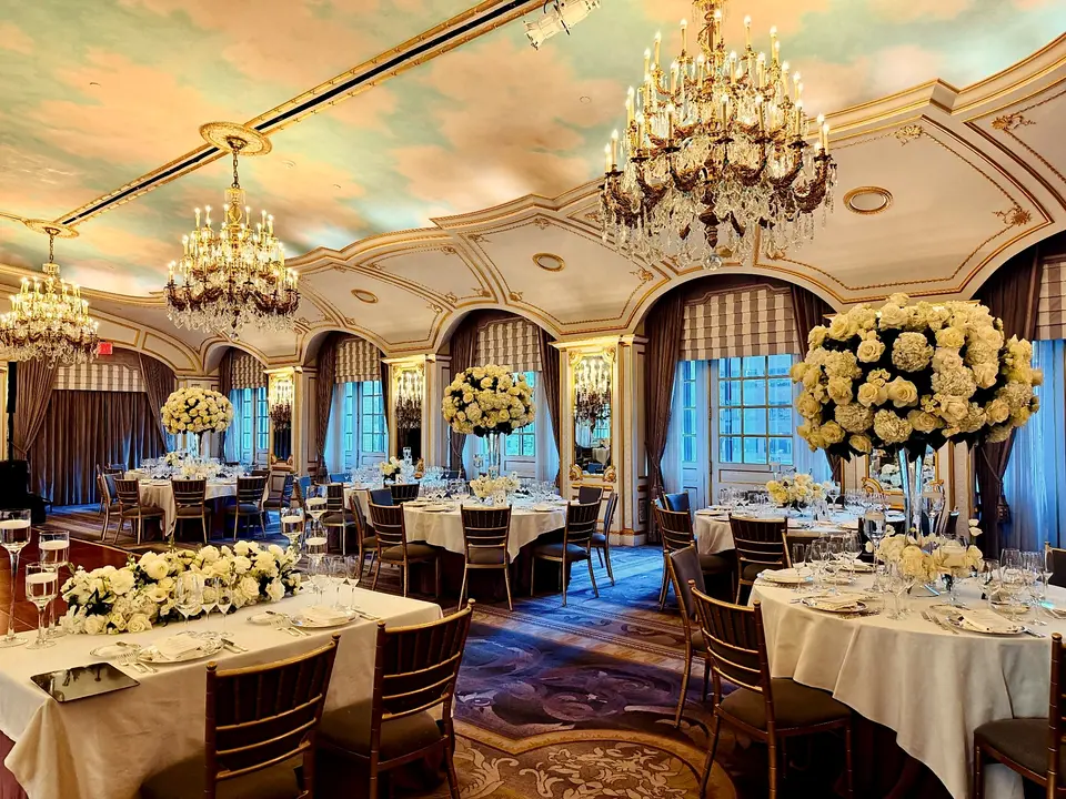 A gilded ballroom with a painted sky ceiling, set for dinner beneath crystal chandeliers, with tall white hydrangea and rose arrangements on every table.