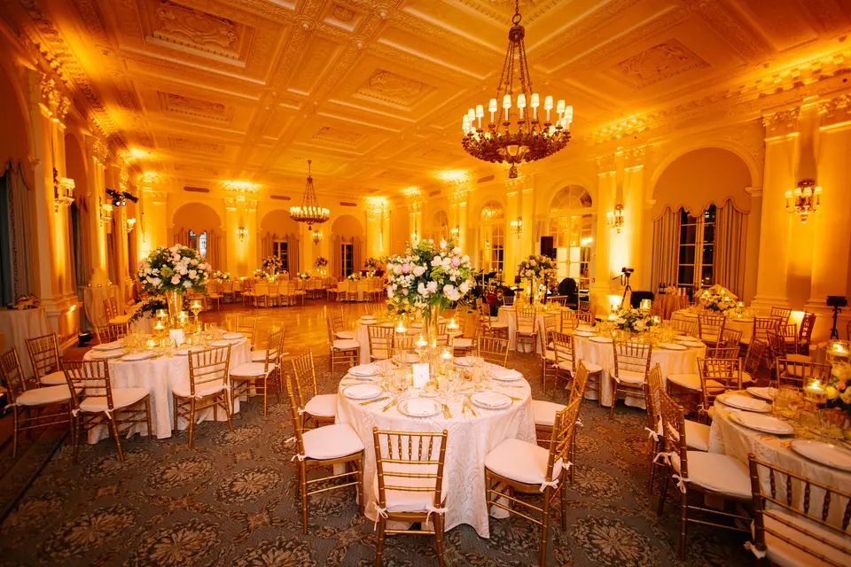 The ballroom in amber light, round tables set with tall white arrangements beneath a great chandelier.