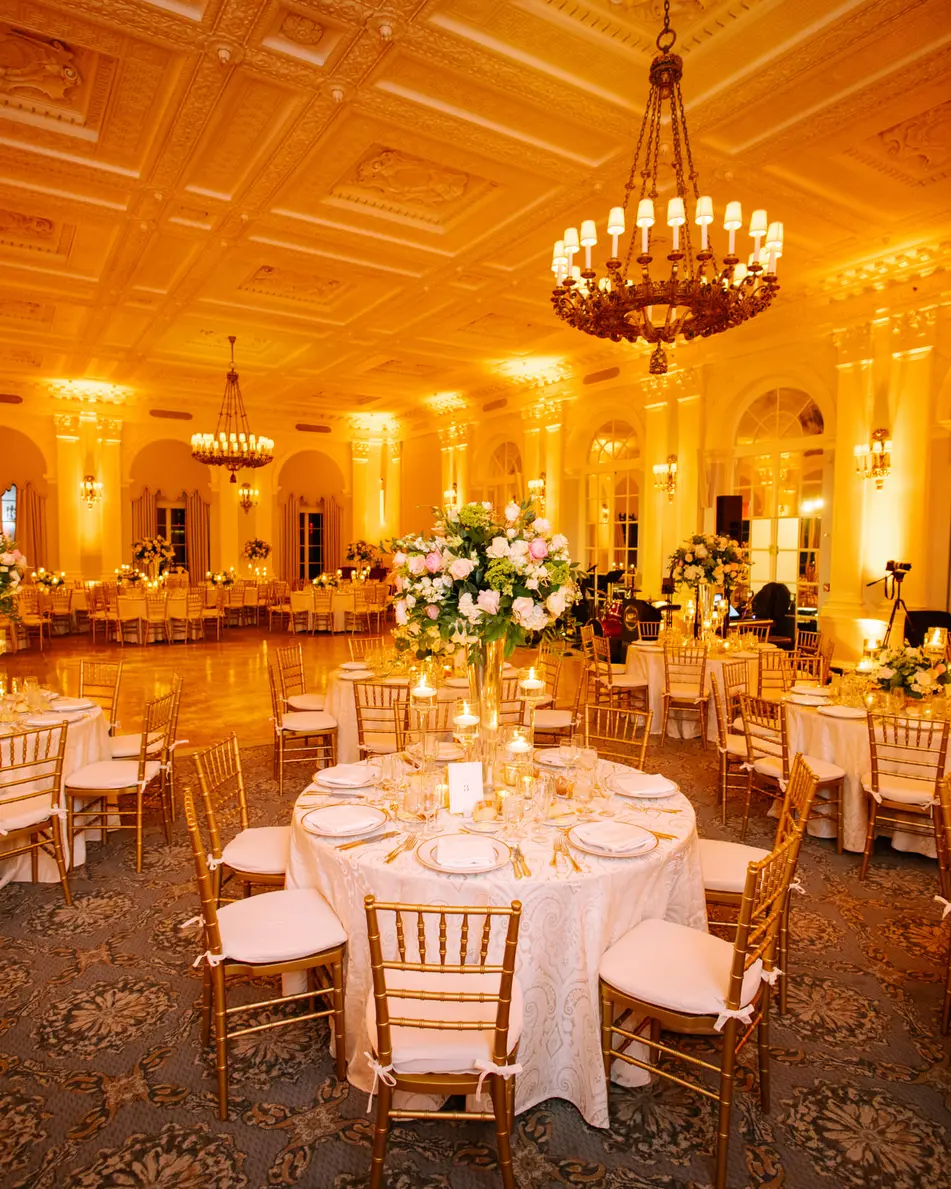 The ballroom in amber light, round tables set with tall white arrangements beneath a great chandelier.