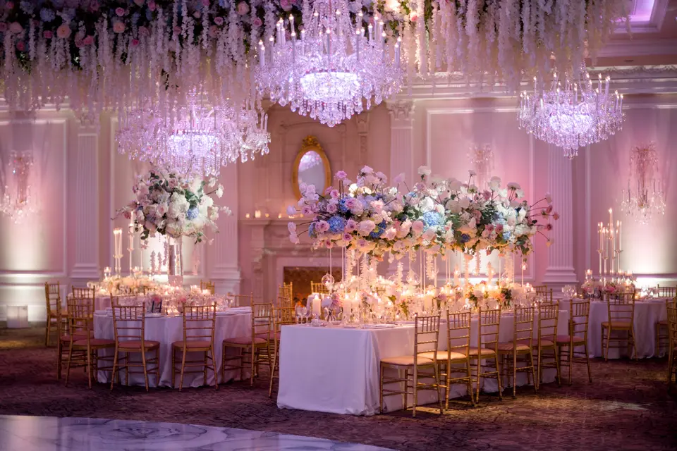 Long tables with gold chairs beneath hanging white flowers and chandeliers, the room washed in soft pink light.
