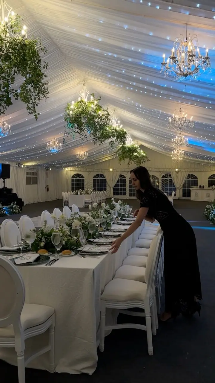 Inside a white tent at Lyndhurst, a member of the team lays a long table set with white chairs and greenery, chandeliers hanging from the draped ceiling.