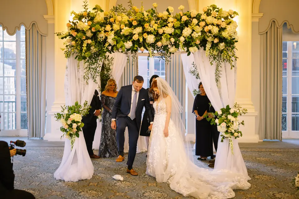 Beneath the flowered chuppah, the groom steps forward as the bride smiles beside him, family standing close.