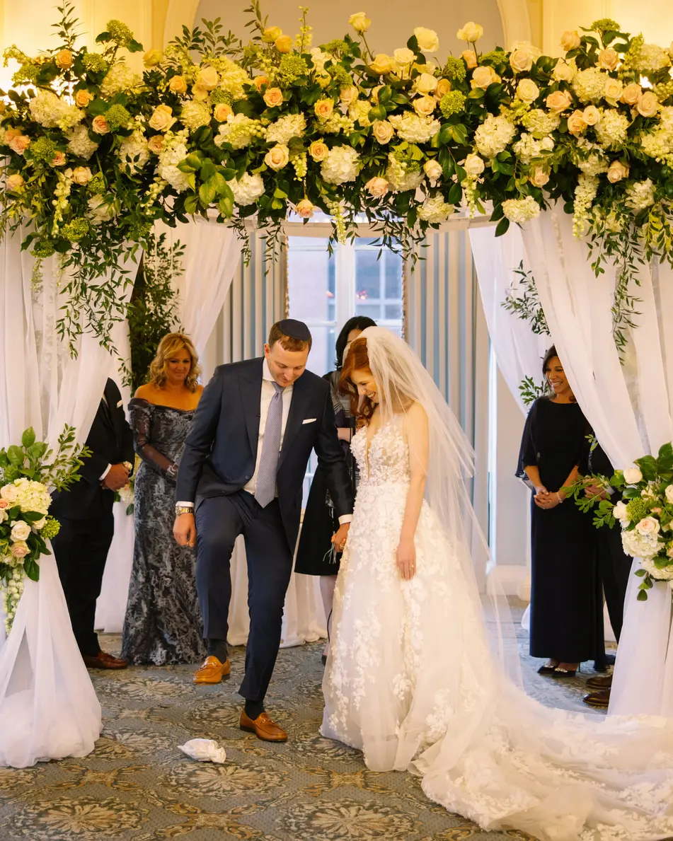 Beneath the flowered chuppah, the groom steps forward as the bride smiles beside him, family standing close.