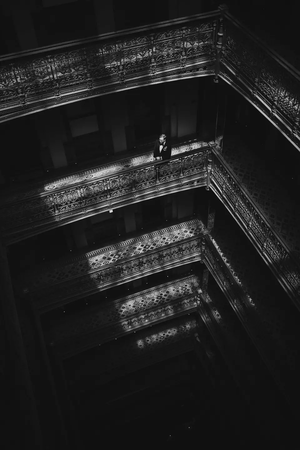 Looking up through a tall atrium of ornate iron balconies, the groom standing alone at a railing high above. Black and white.