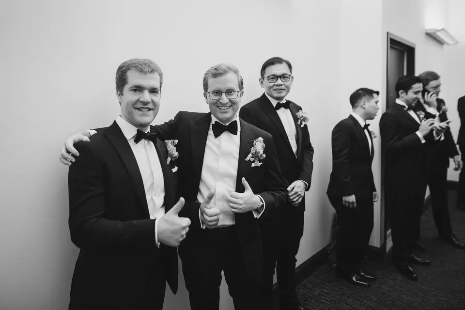 The groom and his groomsmen in tuxedos, two of them grinning and giving a thumbs up. Black and white.