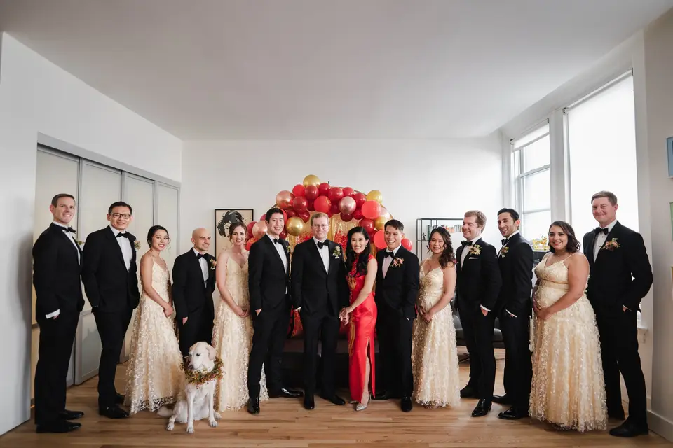 The wedding party lined up in a bright room, the bride in red and the bridesmaids in gold, a dog in a flower collar at their feet and red and gold balloons behind.