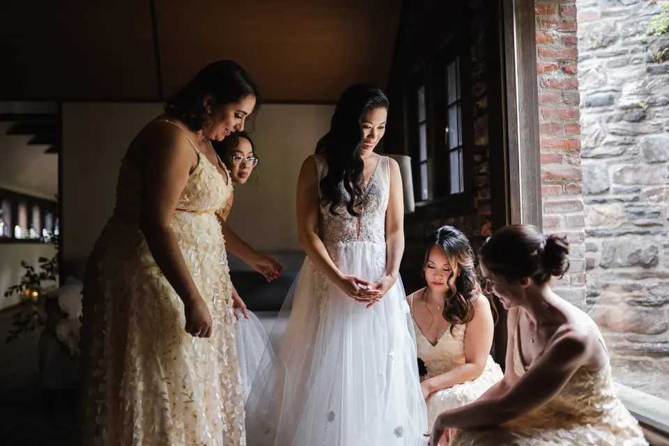 By a window in a stone-walled room, the bridesmaids in gold help the bride with her gown.