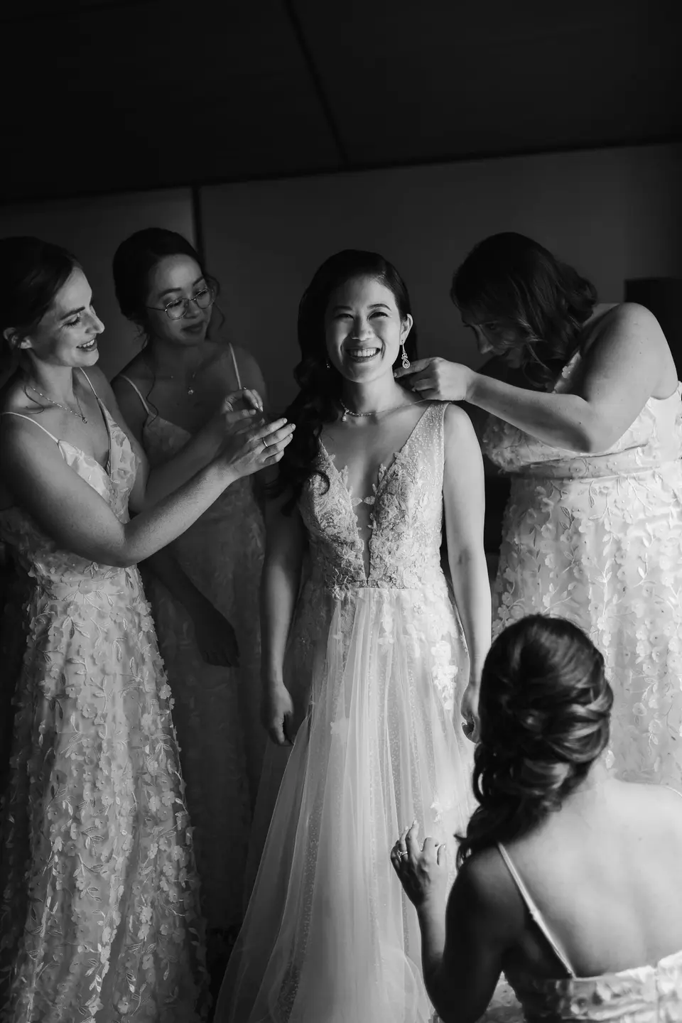 The bridesmaids fasten the bride's necklace as she laughs, one kneeling to straighten her gown. Black and white.