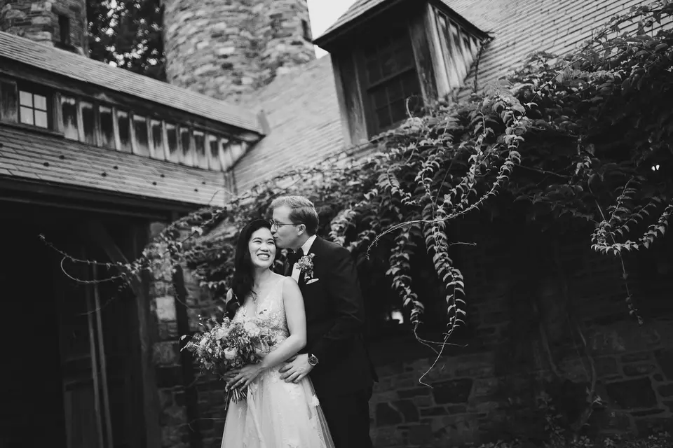 The groom kisses the bride's cheek beside an ivy-covered stone building. Black and white.