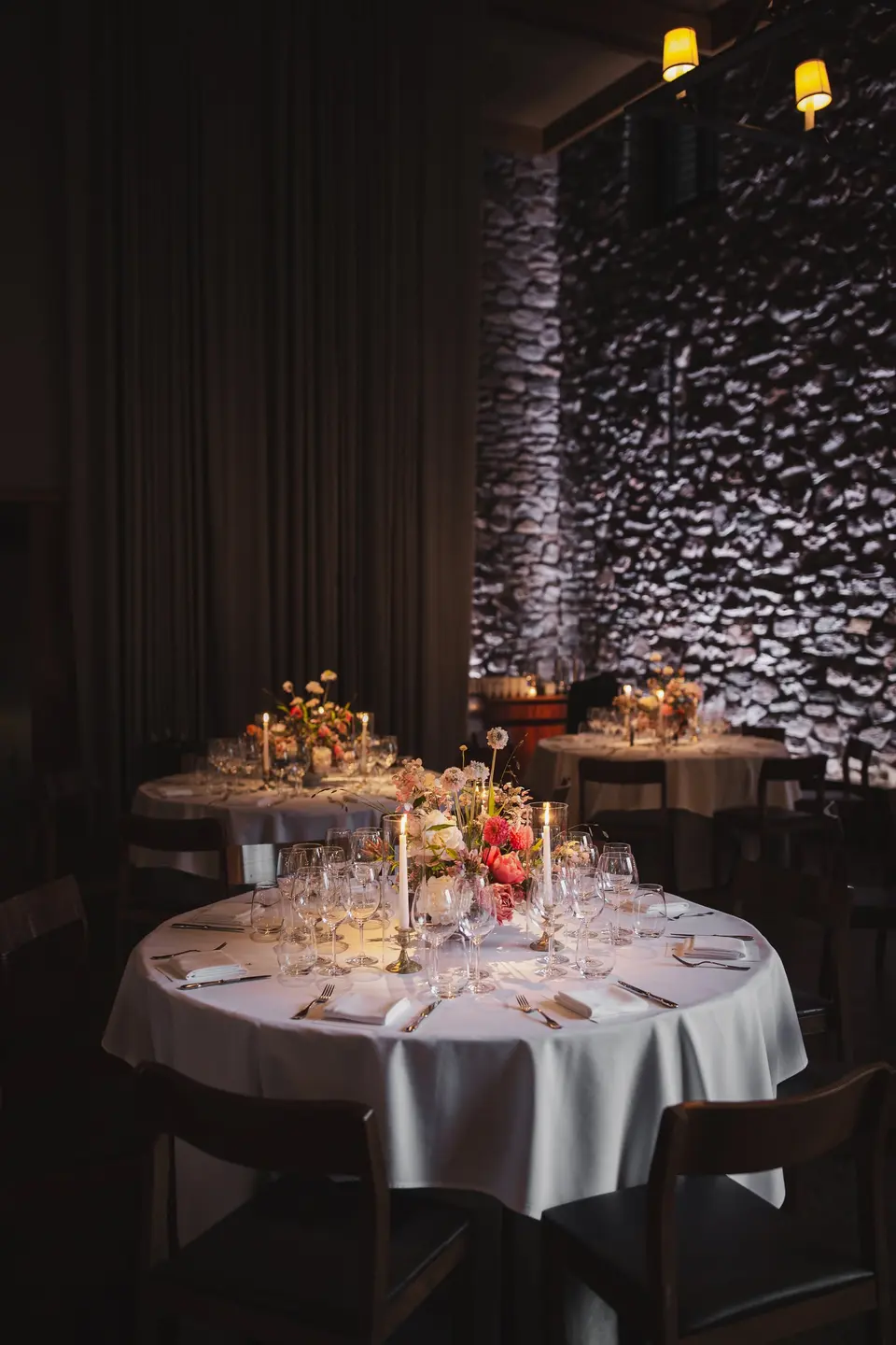 A round table in a dim stone-walled room, set with candles and a low arrangement of orange and pink flowers.