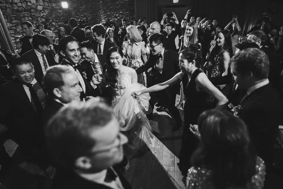 Guests crowd around the bride on the dance floor, laughing and cheering. Black and white.