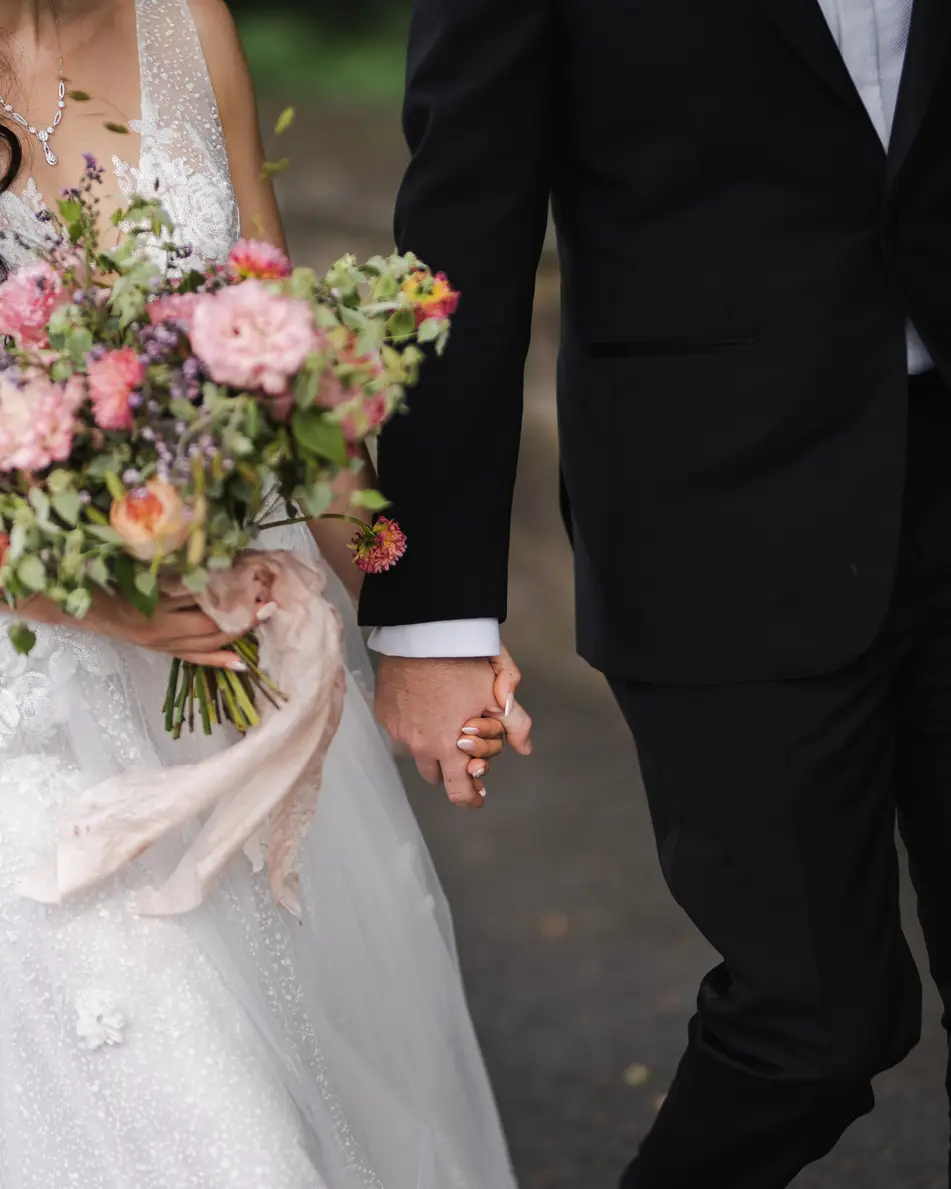The bride holds a loose bouquet of pink roses and trailing ribbon, her hand in the groom's.
