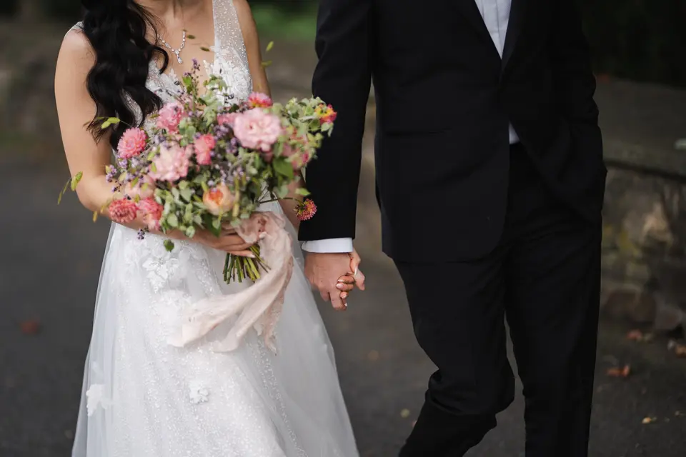 The bride holds a loose bouquet of pink roses and trailing ribbon, her hand in the groom's.