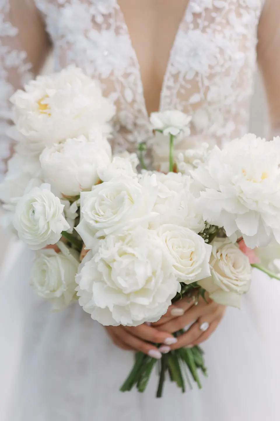 The bride holds a bouquet of white peonies and roses against the lace of her gown.