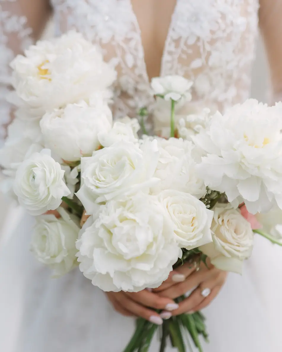 The bride holds a bouquet of white peonies and roses against the lace of her gown.