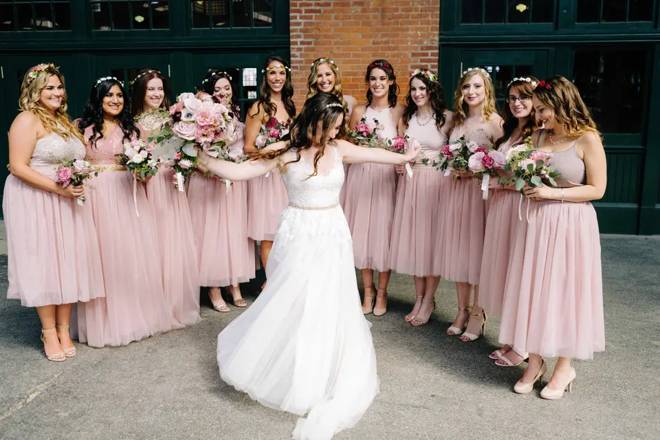 The bride spins with her arms out before her bridesmaids in blush, beneath the terminal canopy.