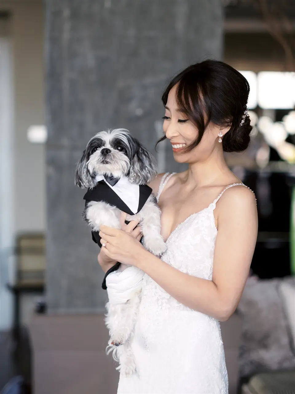 The bride in her gown smiles down at a small black and white dog in a tiny tuxedo, held in her arms.