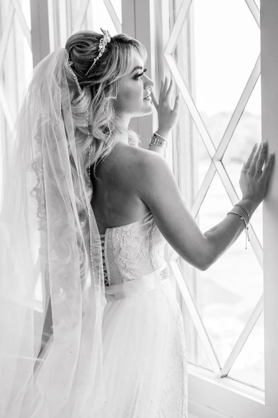 The bride stands at a tall window in her veil, one hand on the frame. Black and white.