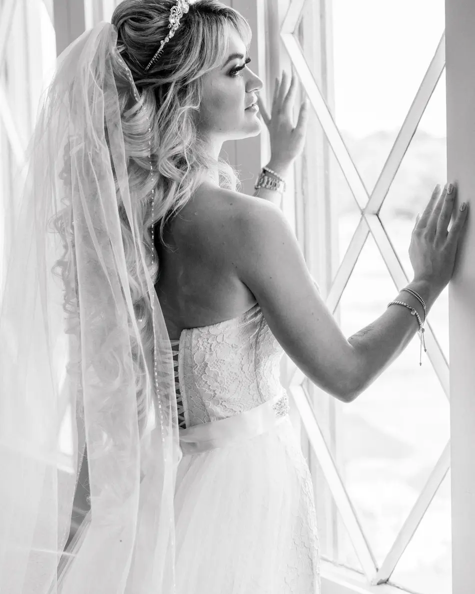 The bride stands at a tall window in her veil, one hand on the frame. Black and white.