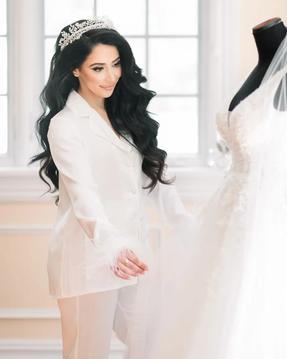 The bride, in a white robe and a crystal tiara, stands by a window beside her gown.