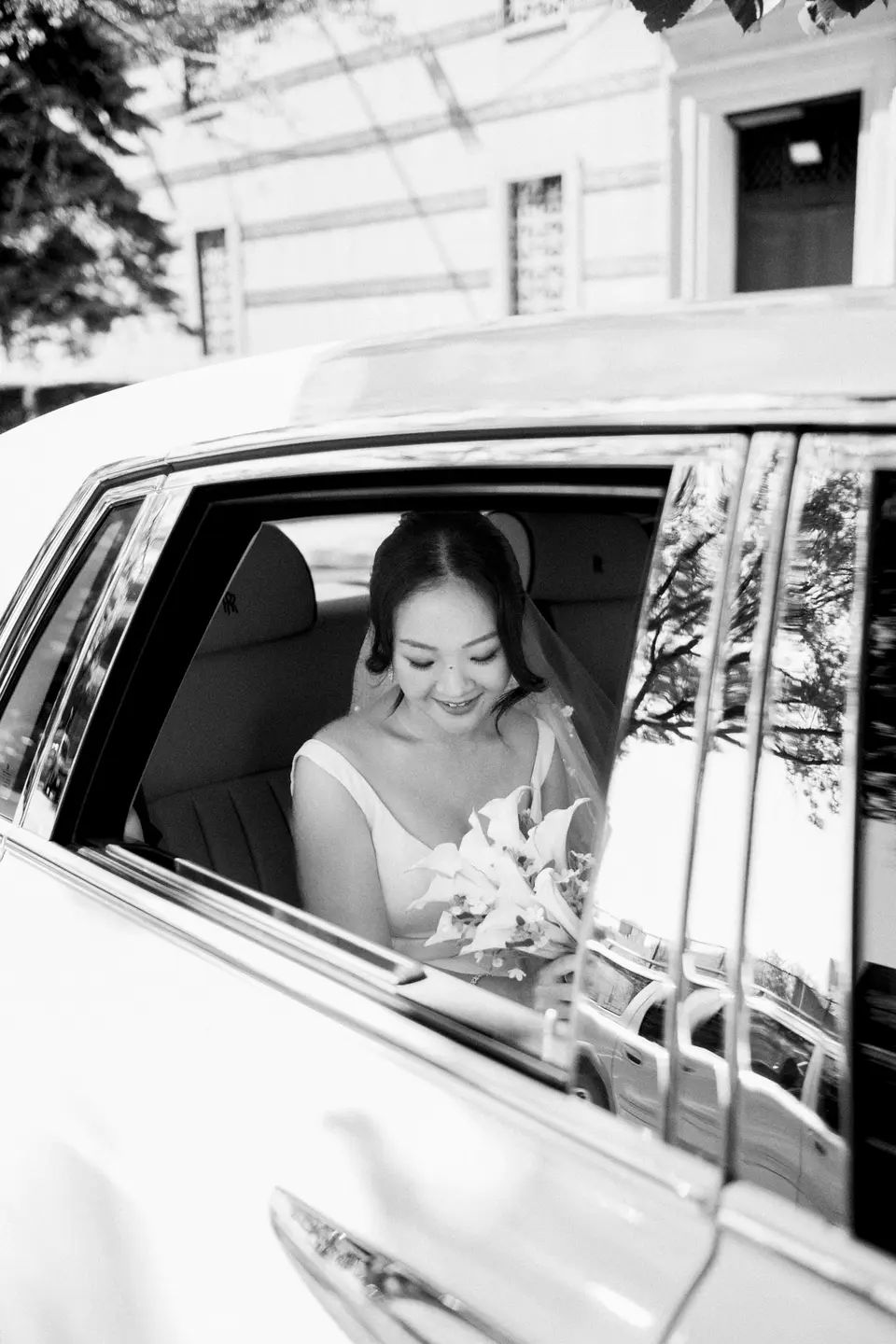 The bride looks out from the window of a white car, smiling, her bouquet in her lap. Black and white.