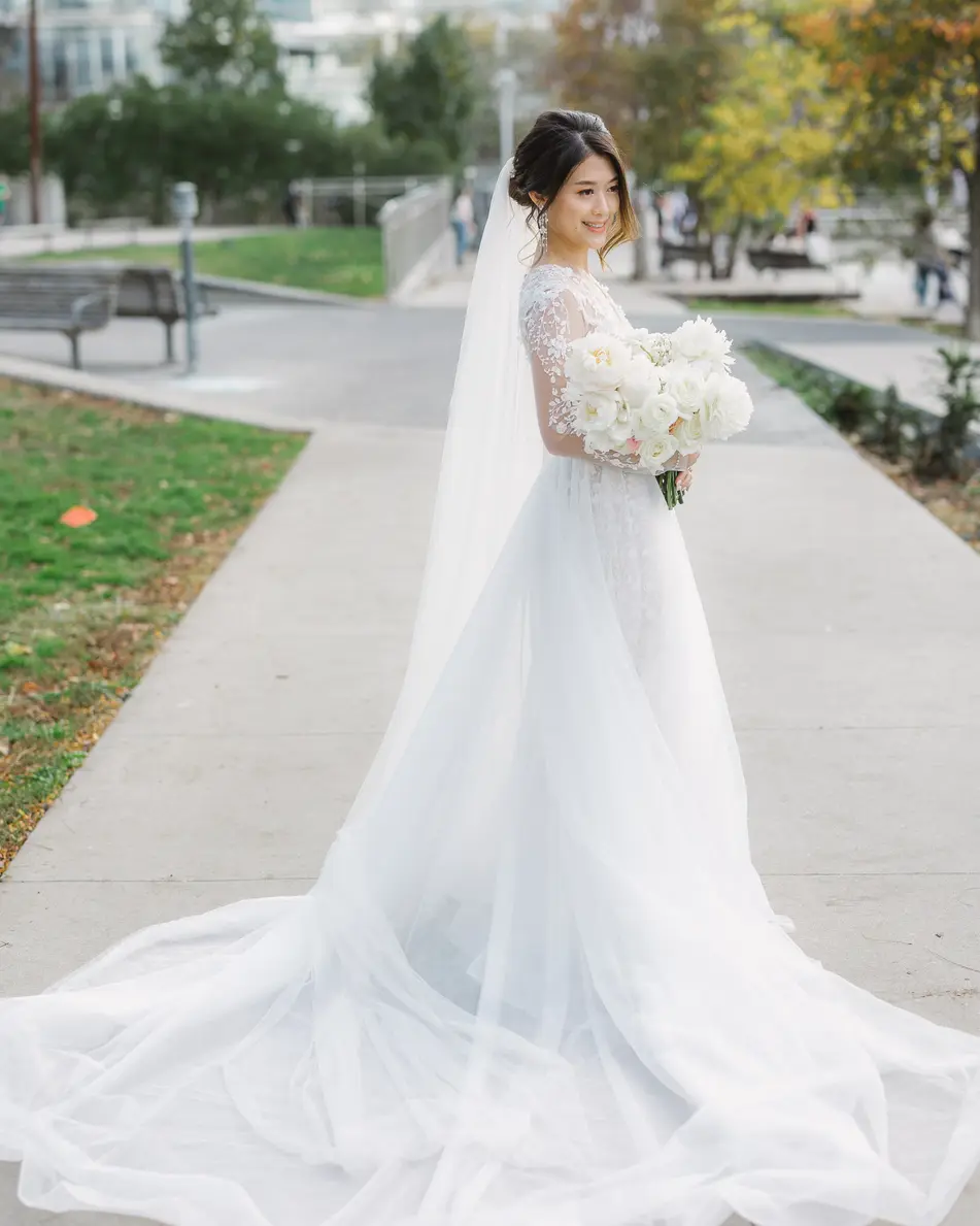 The bride stands on a park path in autumn, her long veil and train spread across the paving.
