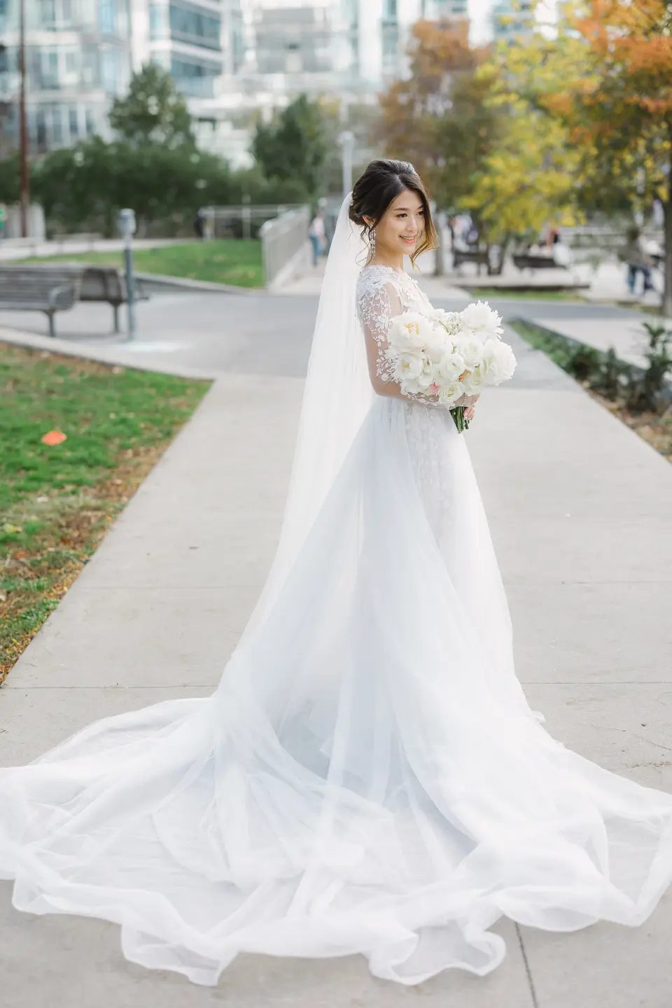 The bride stands on a park path in autumn, her long veil and train spread across the paving.