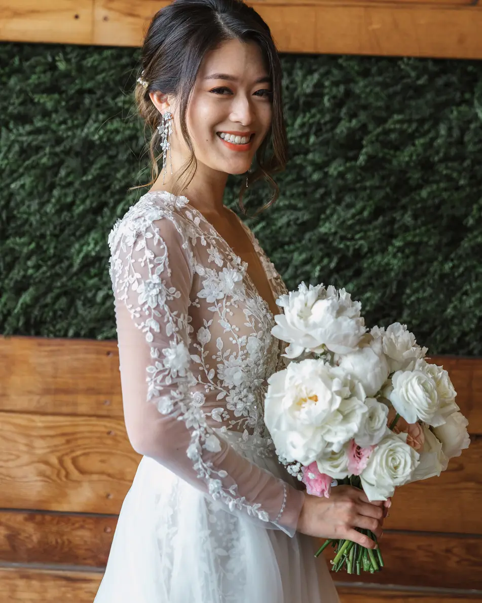 The bride smiles over her shoulder, holding white peonies and roses, in a lace gown with sheer sleeves against a wall of wood and greenery.
