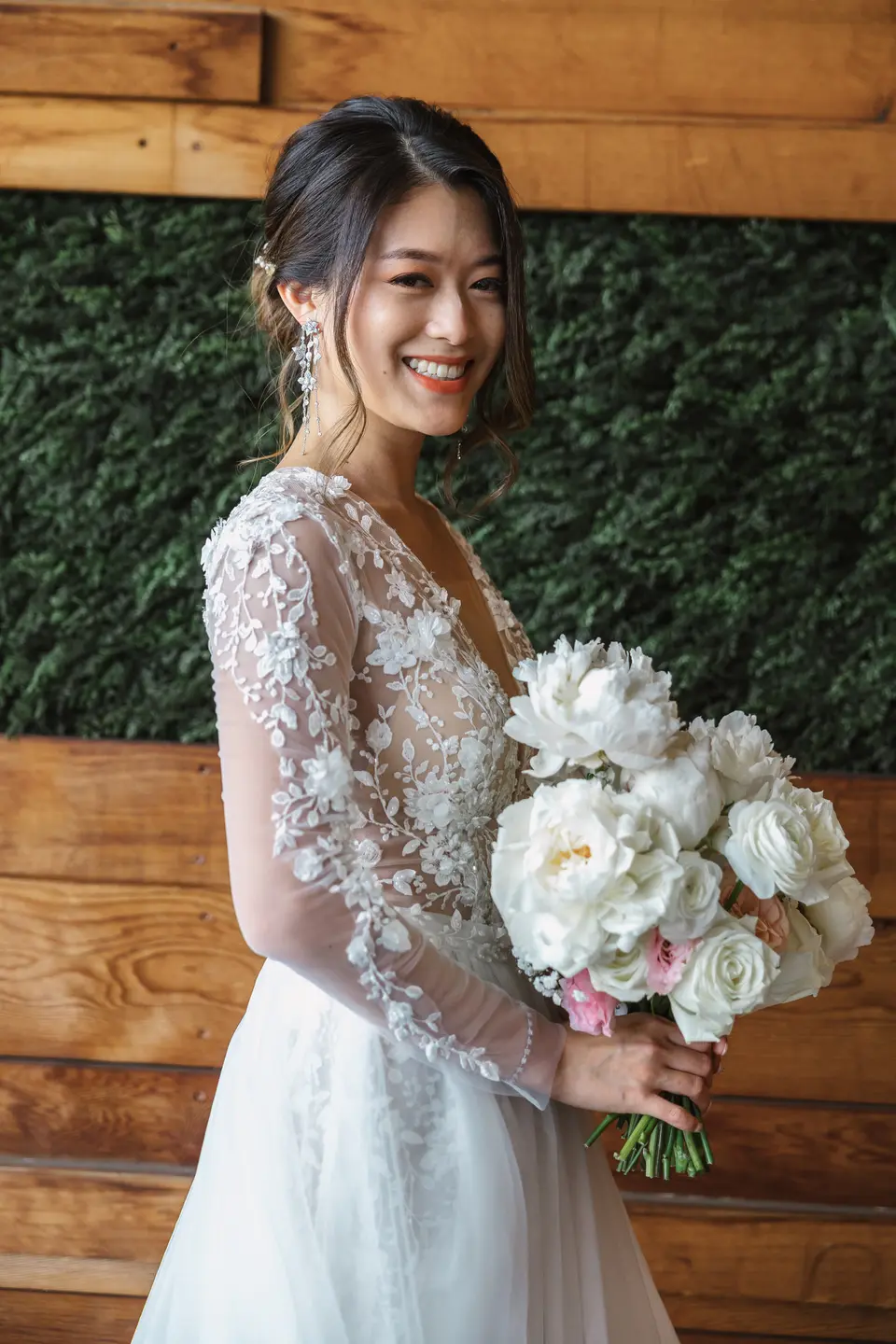 The bride smiles over her shoulder, holding white peonies and roses, in a lace gown with sheer sleeves against a wall of wood and greenery.