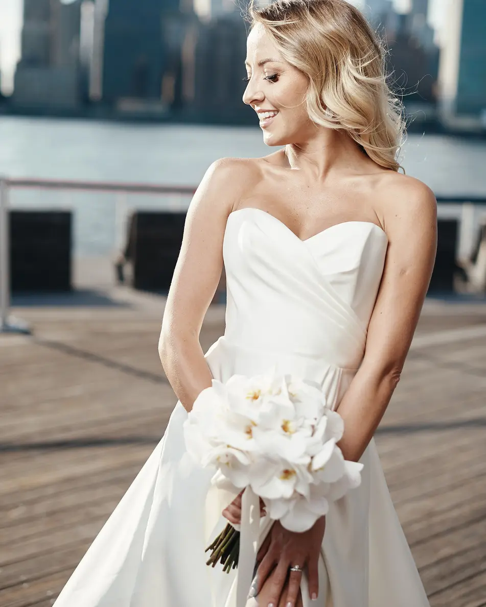The bride stands on a wooden pier holding white orchids, looking down and smiling, the skyline across the water.