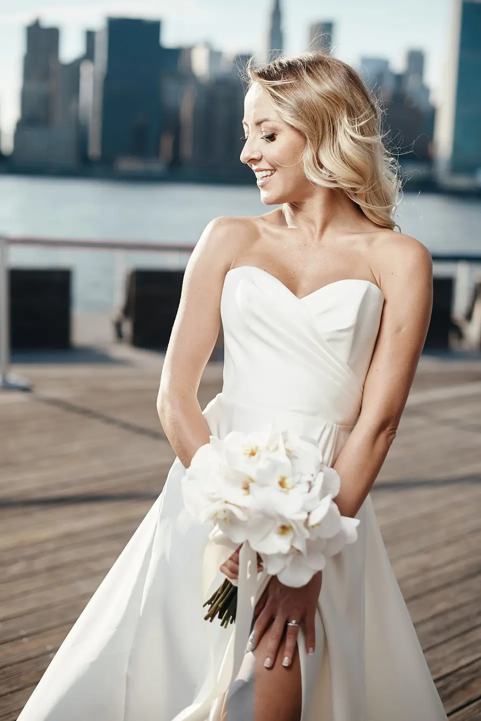 The bride on a wooden pier holding white orchids, the Manhattan skyline across the water.