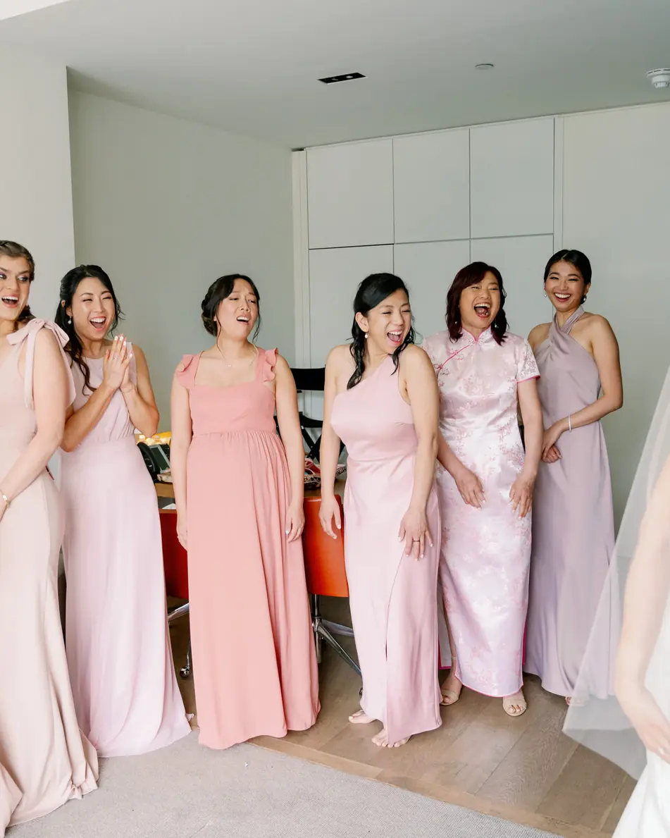 Bridesmaids in shades of pink and lilac see the bride in her gown and veil for the first time, hands to their faces.