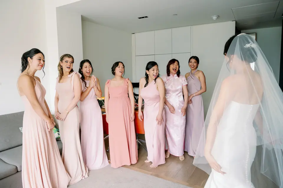 Bridesmaids in shades of pink and lilac see the bride in her gown and veil for the first time, hands to their faces.