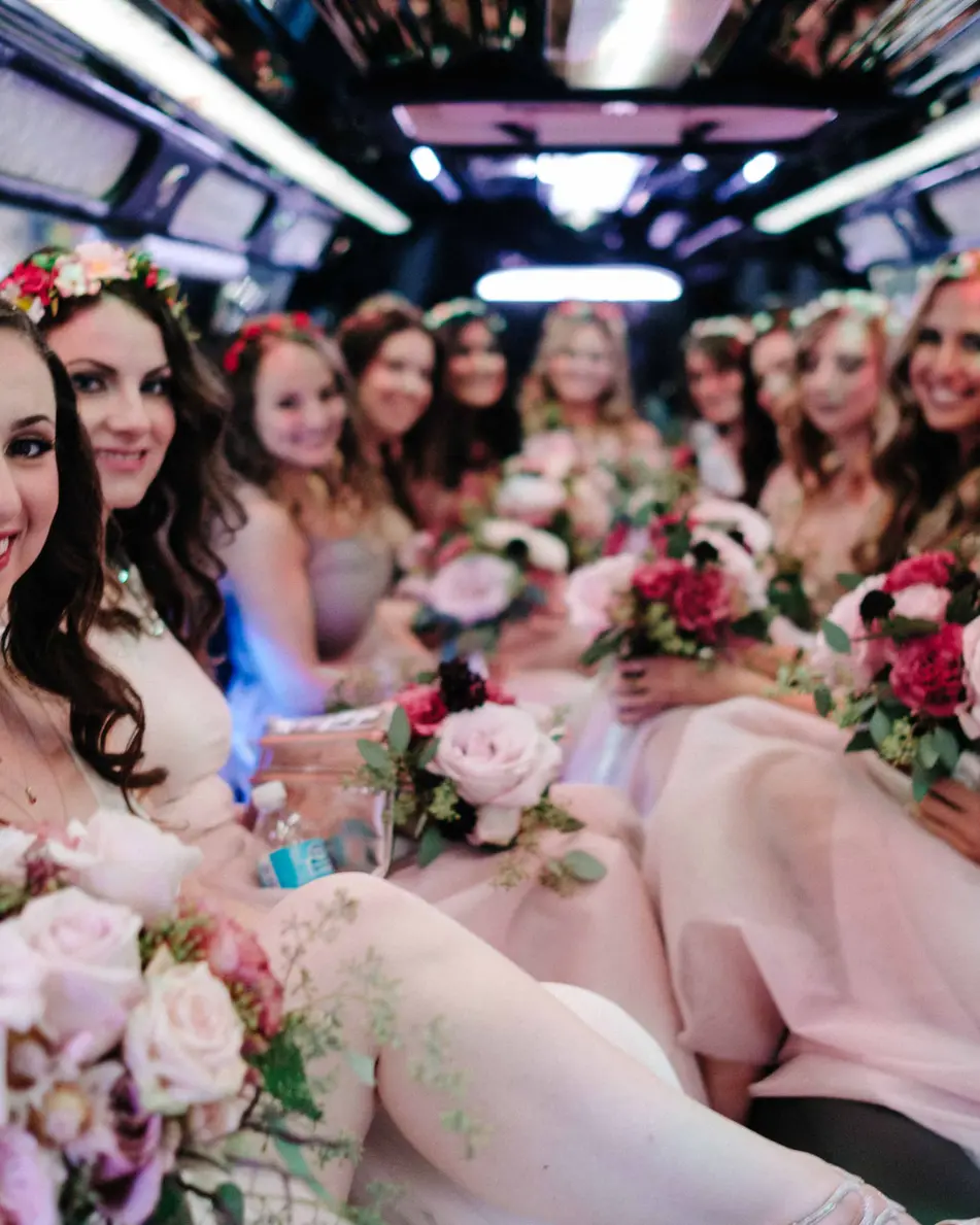 The bride and her bridesmaids, in flower crowns, laugh together inside a limousine.