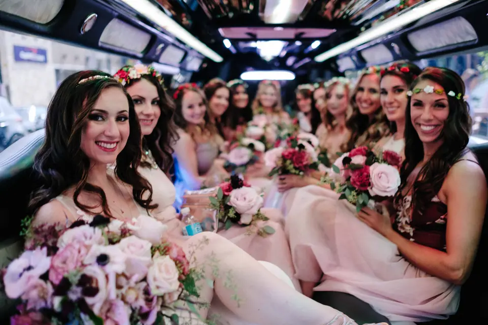 The bride and her bridesmaids, in flower crowns, laugh together inside a limousine.