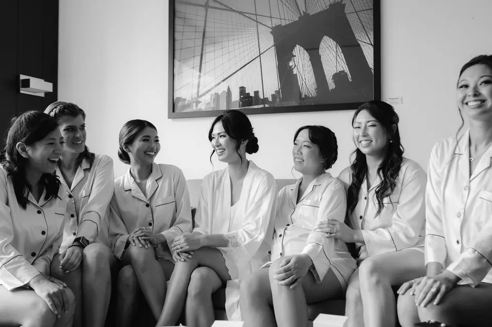 The bride and her bridesmaids in matching white pajamas, laughing together on a sofa. Black and white.
